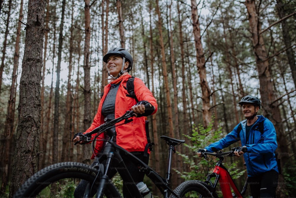A low angle view of senior couple bikers walking and pushing e-bikes outdoors in forest in autumn day.