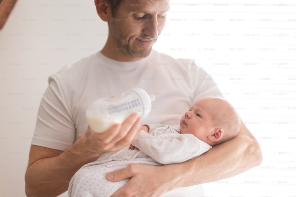 A father holding and feeding his newborn son with milk bottle at home.