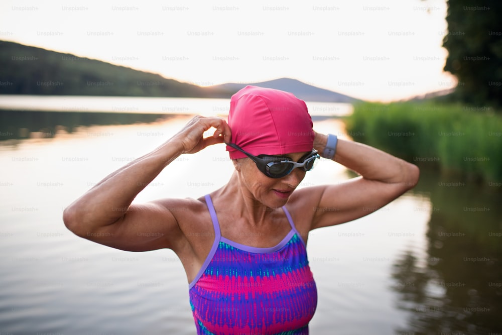 A portrait of active senior woman swimmer standing and putting on goggles outdoors by lake.