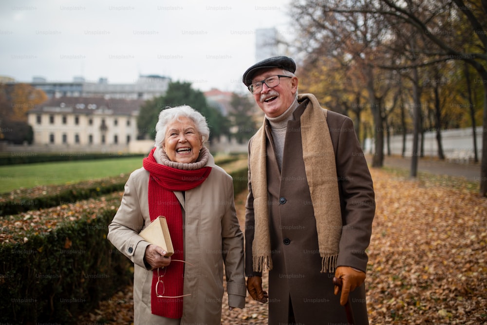 A happy senior couple on walk outdoors in park in autumn, embracing, laughing and looking at camera