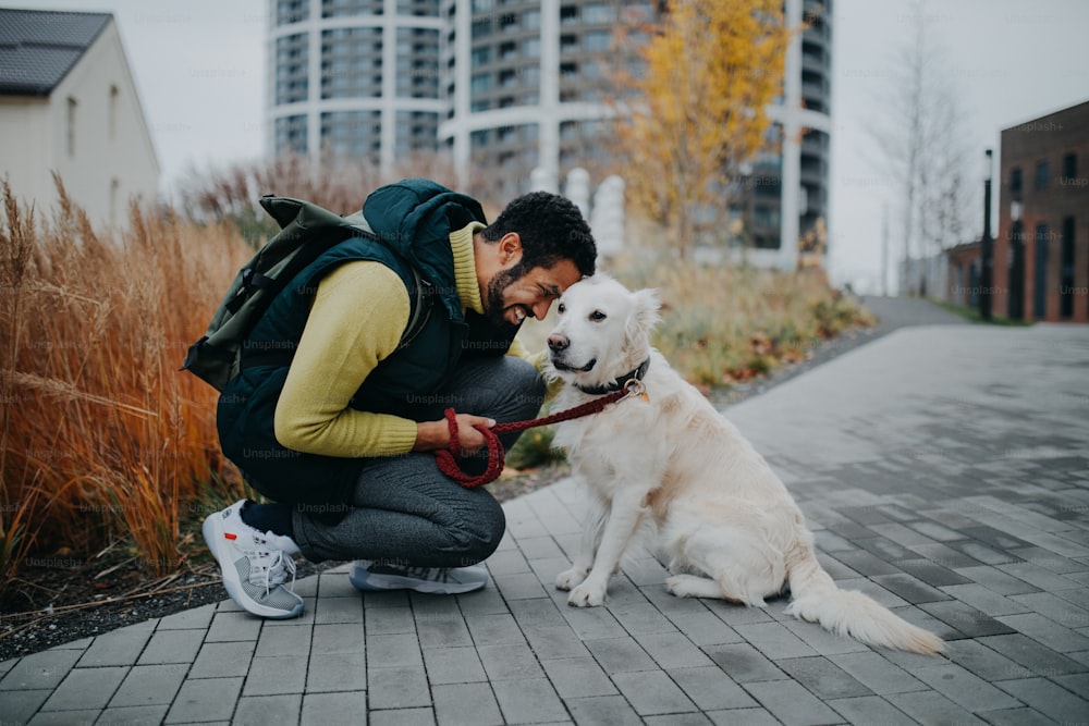 A happy young man squatting and embracing his dog during walk outdoors in city.
