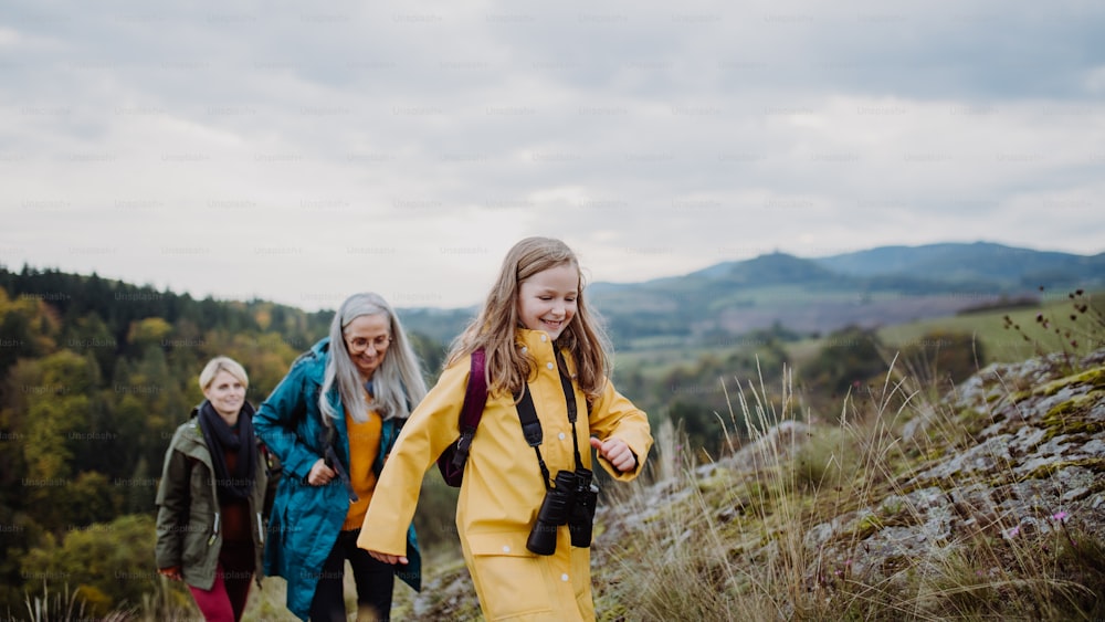 A portrait of small girl with mother and grandmother standing and looking at camera outoors in nature.A rear view of small girl with mother and grandmother hiking outoors in autumn nature.