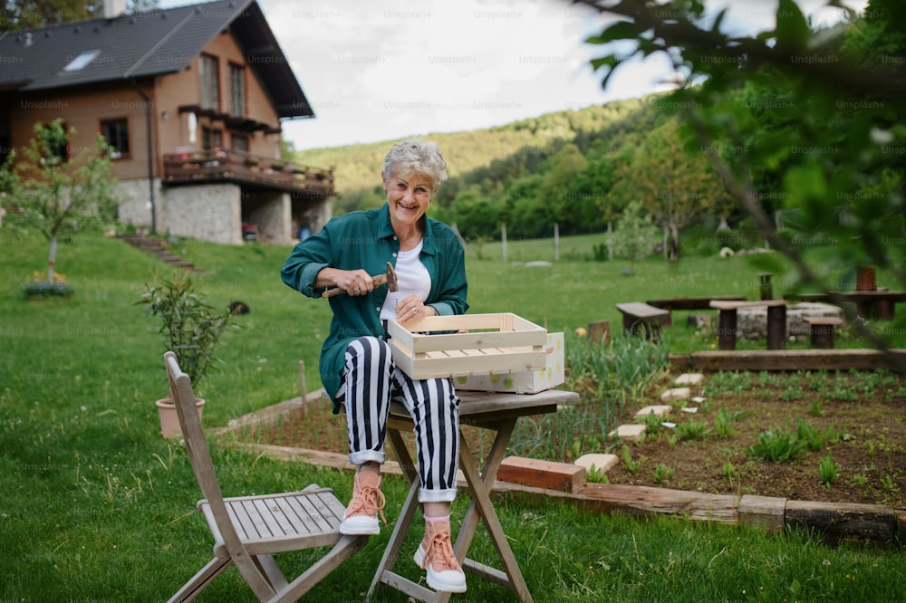 A happy senior woman renovating wooden crate outdoors in garden, looking at camera.