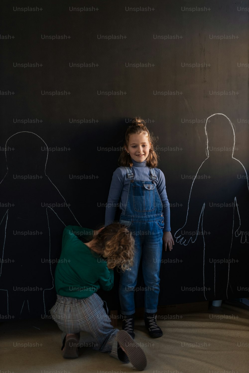 A rear view of little girl drawing her friends with chalks on blackboard wall indoors in playroom.