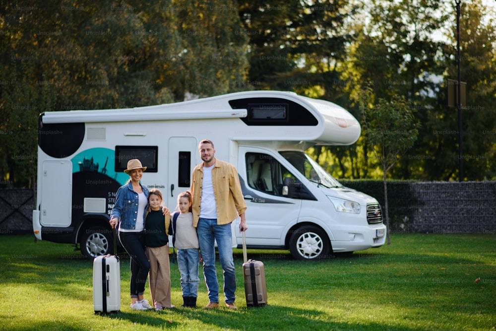 A happy young family with luggage looking at camera with camper at background outdoors in garden.