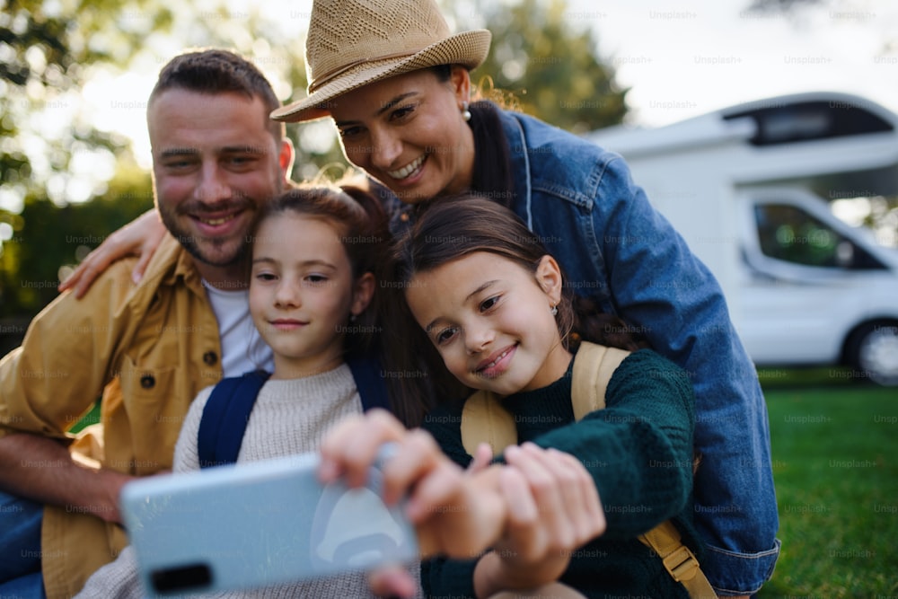 Une jeune famille heureuse avec deux enfants prenant un selfie avec une caravane à l’arrière-plan à l’extérieur.