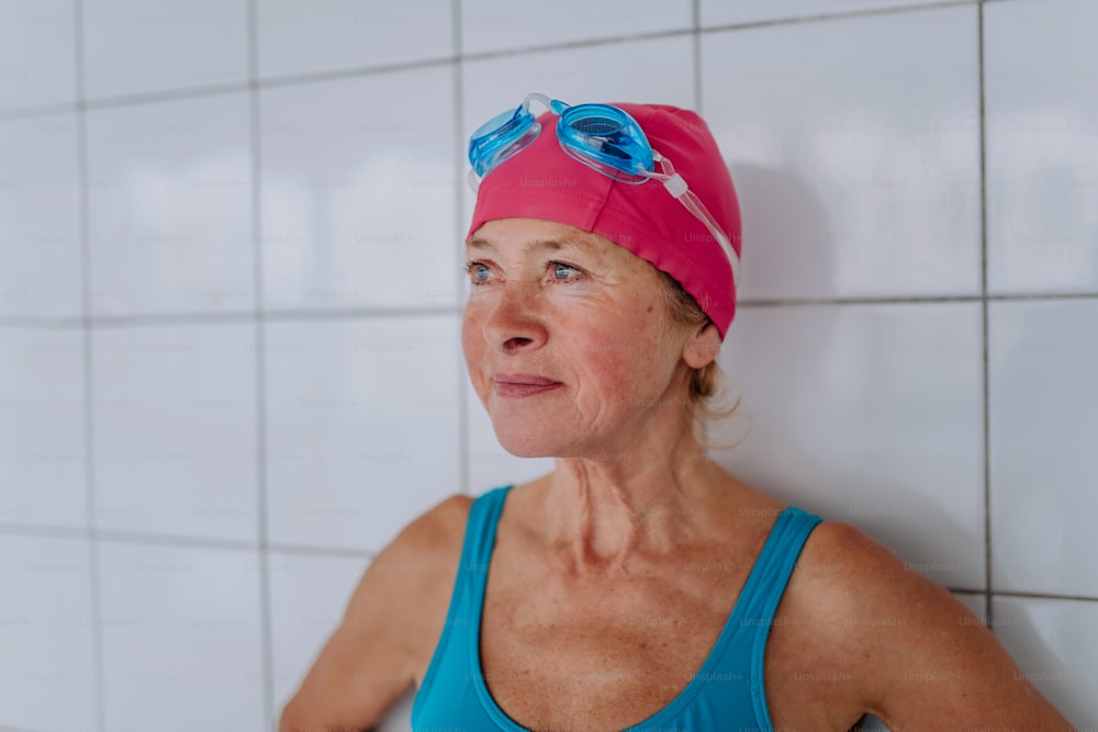 An active senior swimmer woman standing against white tiles wall indoors in swimming pool.