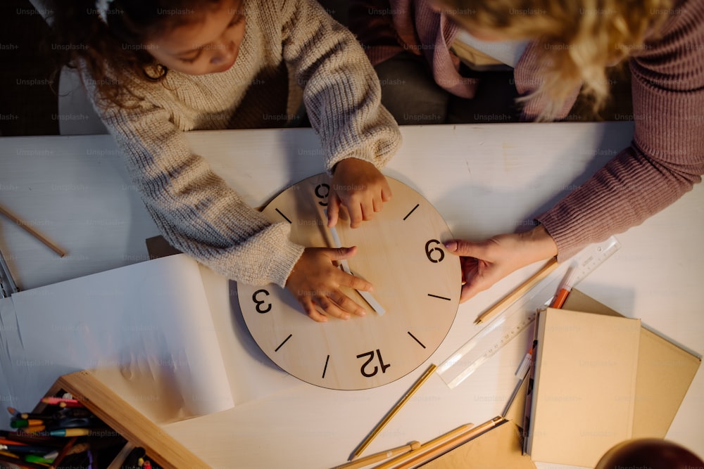 Una vista dall'alto di angolo della bambina che impara il tempo con l'orologio di legno con sua madre la sera a casa.