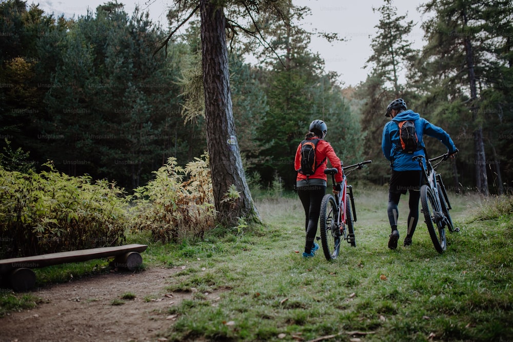 Una vista trasera de una pareja de ciclistas mayores caminando y empujando bicicletas eléctricas al aire libre en el bosque en un día de otoño.
