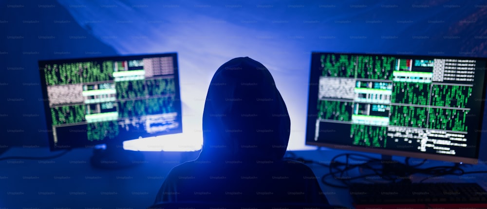 Eine Rückansicht eines vermummten Hackers per Computer in der Dunkelkammer in der Nacht, Cyberwar-Konzept.