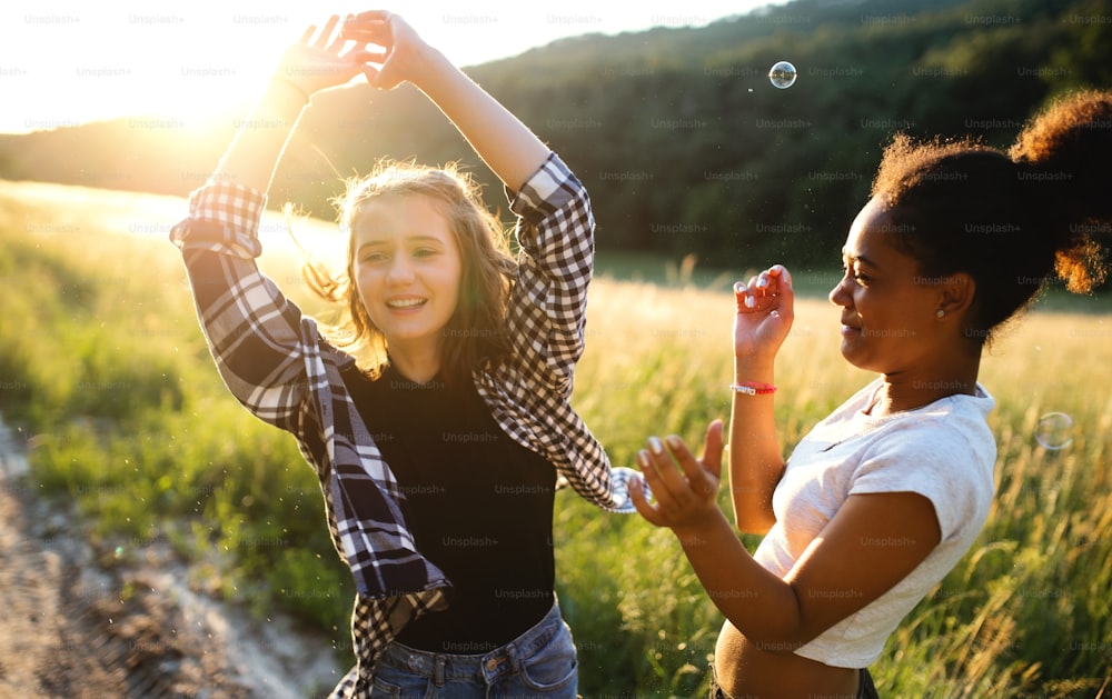 Cheerful young teenager girls friends outdoors in nature at sunset, blowing bubbles.