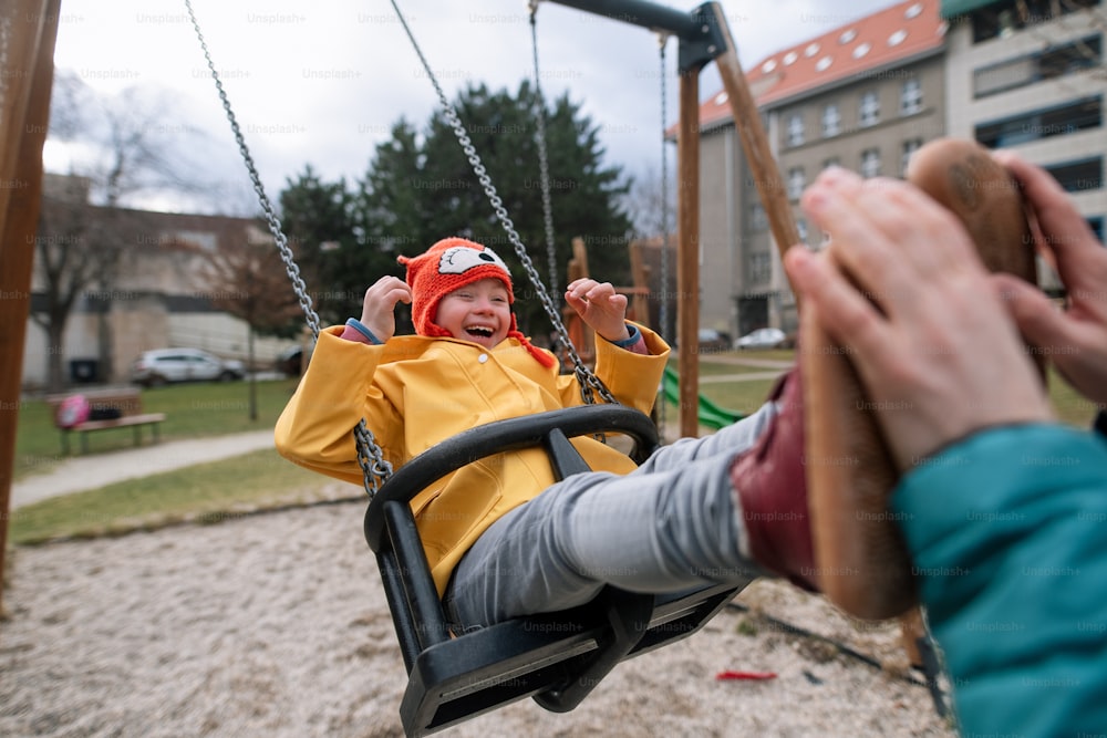 An unrecognizable father pushing his little daughter with Down syndrome on swing outdoors in playgraound.