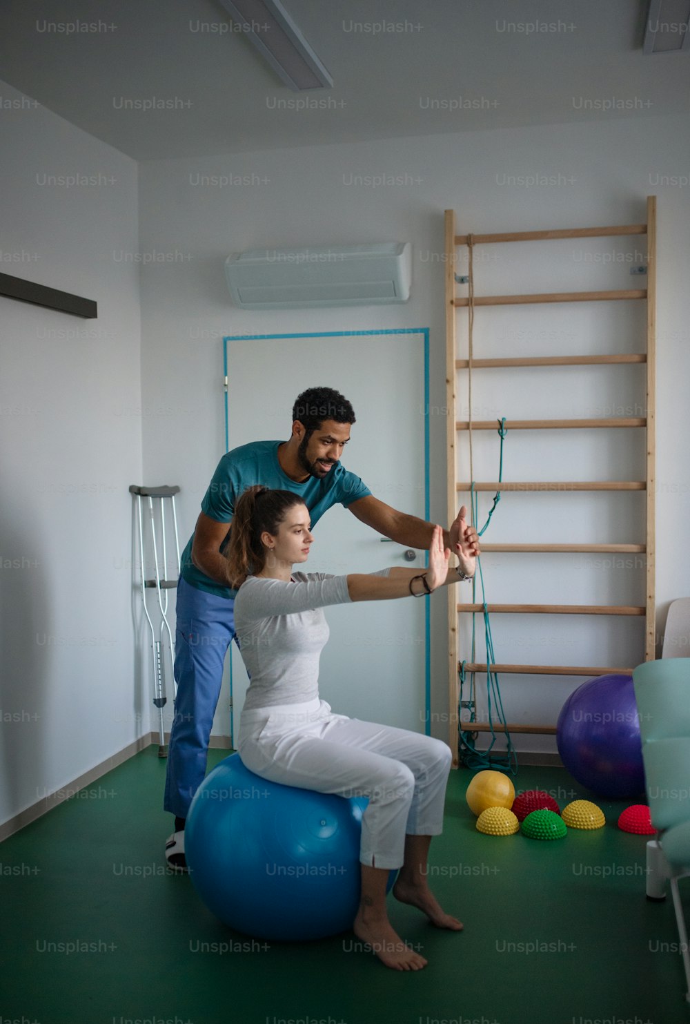 A young male physiotherapist exercising with young woman patient on ball in a physic room