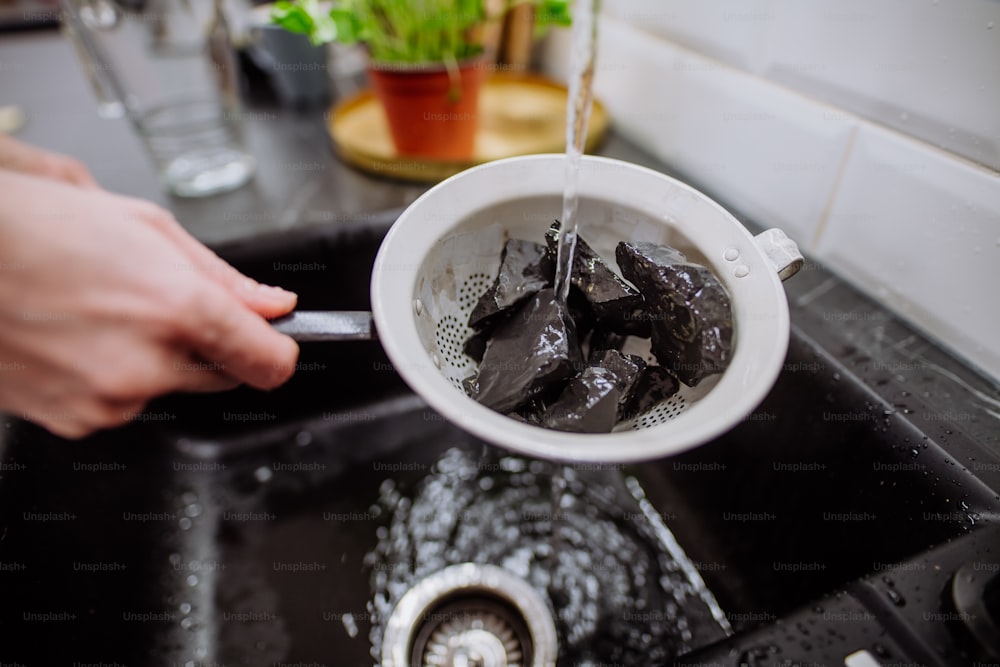 A woman cleaning shungite stones in sieve with pouring water in sink.
