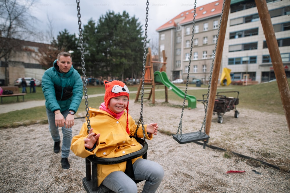 A father pushing his little daughter with Down syndrome on swing outdoors in playgraound.