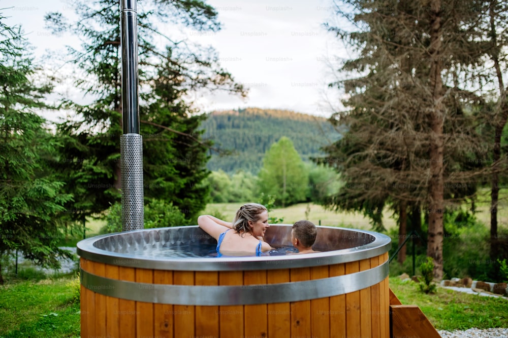 Mother with her little son enjoying bathing in wooden barrel hot tub in a terrace of the cottage. Wooden bathtub with a fireplace to burn wood and heat water. Rear view.
