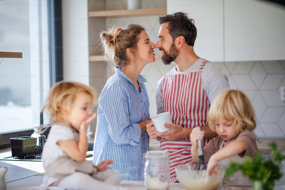 Vista frontal da família jovem com duas crianças pequenas dentro de casa na cozinha, cozinhando.