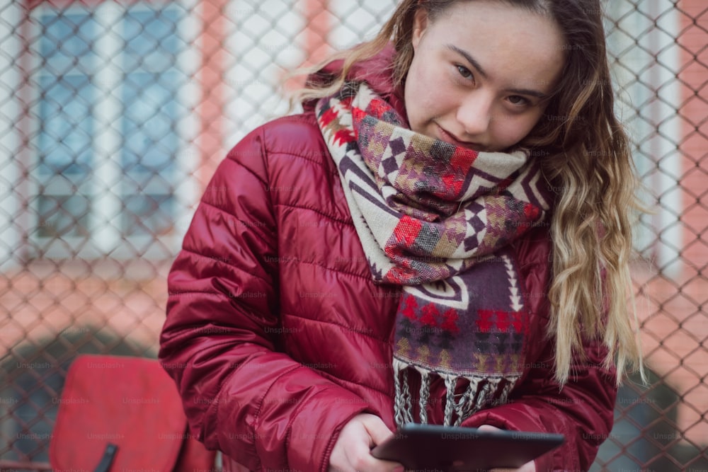 A young woman with Down syndrome using tablet in town in winter and looking at camera.