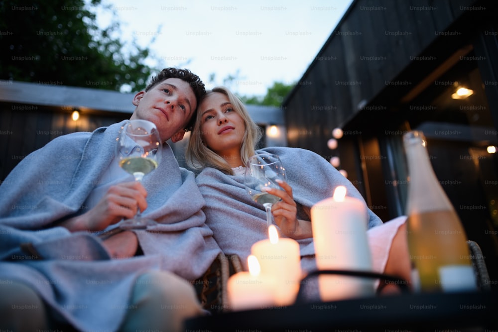 Um jovem casal com vinho descansando ao ar livre no terraço à noite, fim de semana em casa minúscula no campo, vida sustentável.