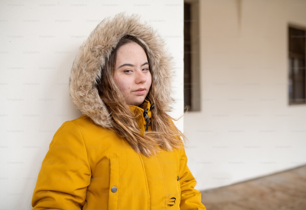A thoughtful young woman with Down syndrome weraing parka, leaning the wall and looking at camera.