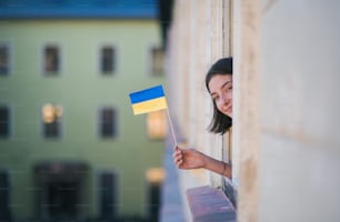 Ein glückliches Mädchen, das die ukrainische Flagge aus dem Fenster ragt und in die Kamera schaut, Solidarität mit der Ukraine im Kriegskonzept.