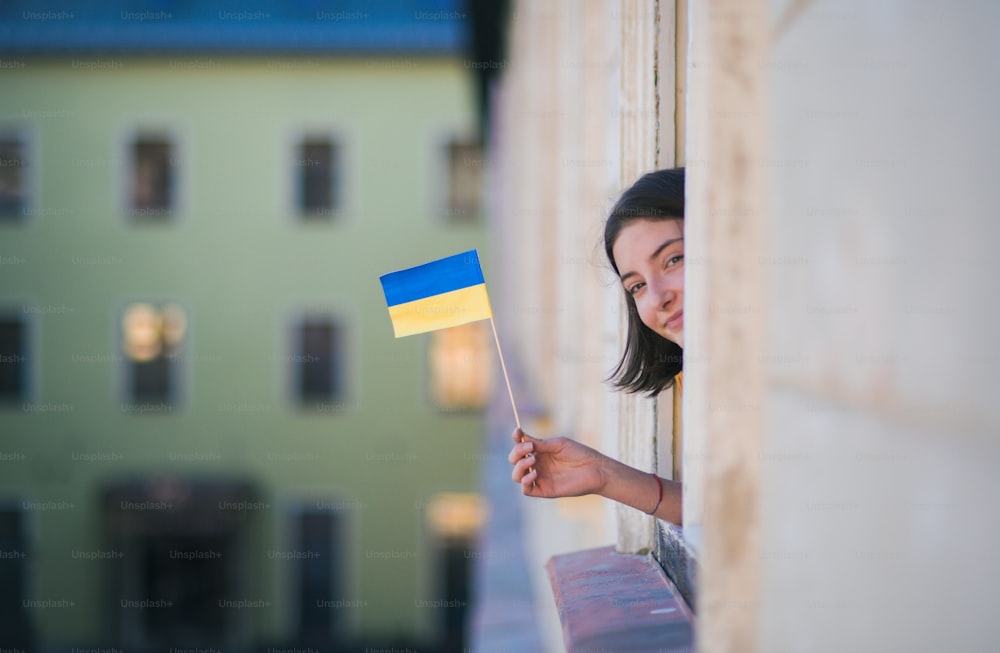 A happy girl sticking Ukrainian flag out from window and looking at camera, solidarity with Ukraine in war concept.
