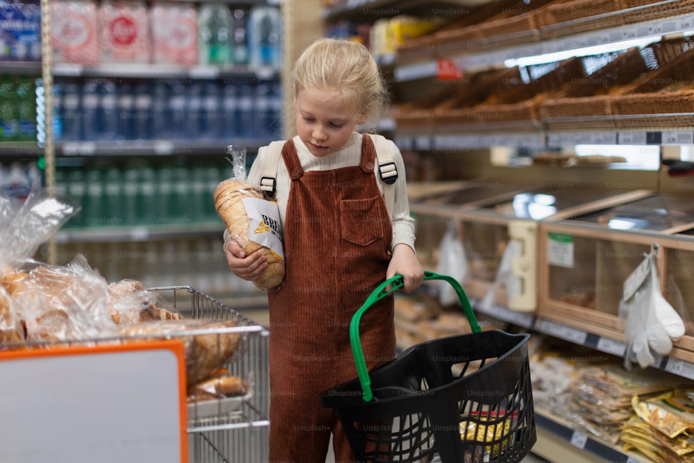 Petite fille debout au milieu du supermarché et achetant du pain, tenant un panier à provisions.