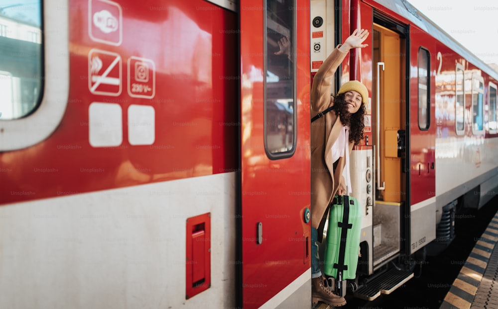 Uma mulher jovem e feliz do viajante com bagagem em pé na porta do trem acenando para fora do trem na plataforma da estação de trem