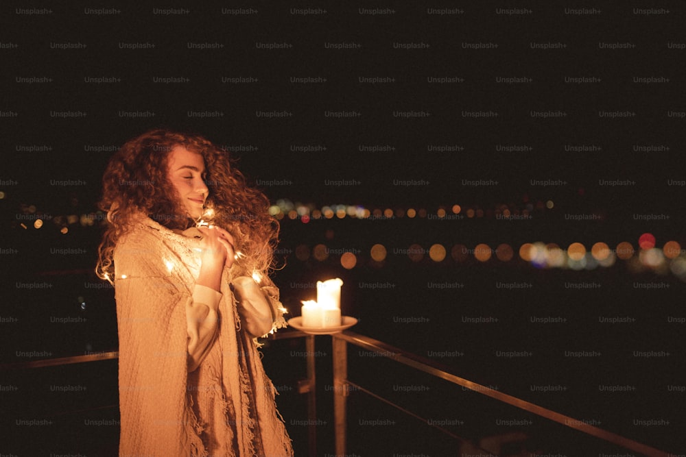 Una joven relajada envuelta en una manta de pie en el balcón con velas y disfrutando del tiempo por la noche, estilo de vida hygge.