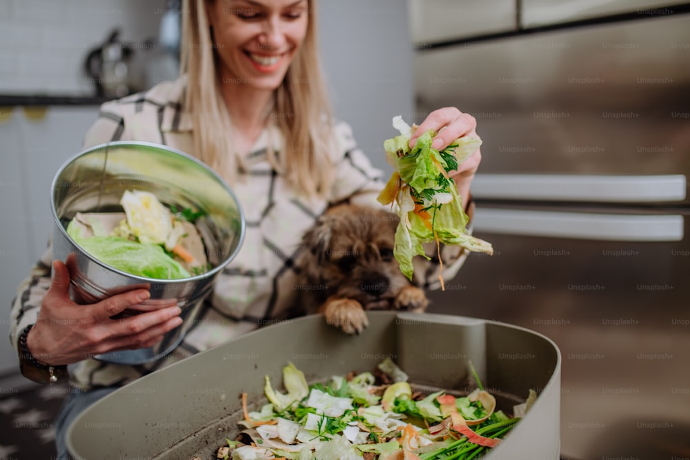 Una donna che getta talee di verdura in un secchio di compost in cucina e dà da mangiare al cane.