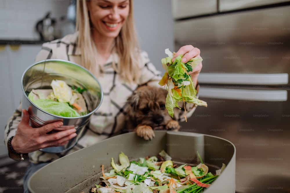 A woman throwing vegetable cuttings in a compost bucket in kitchen and feeding dog.