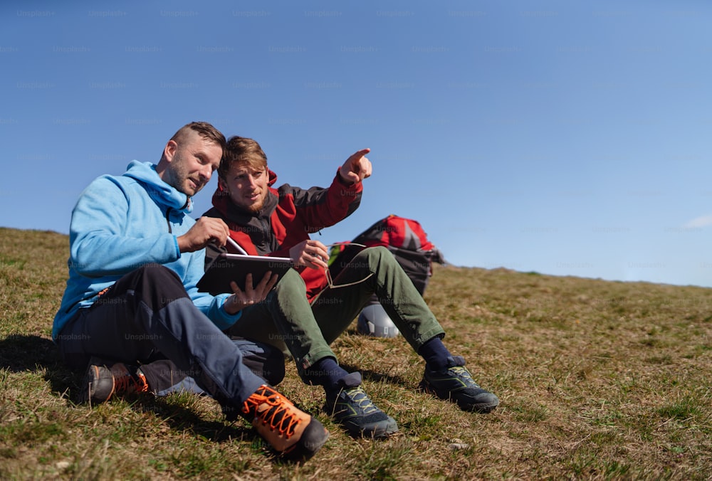 Two paragliders men sitting on the top of the mountain with tablet and looking at view.