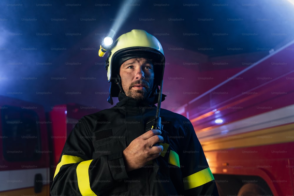 Uma visão de baixo ângulo do bombeiro m conversando com walkie talkie com caminhão de bombeiros ao fundo à noite.