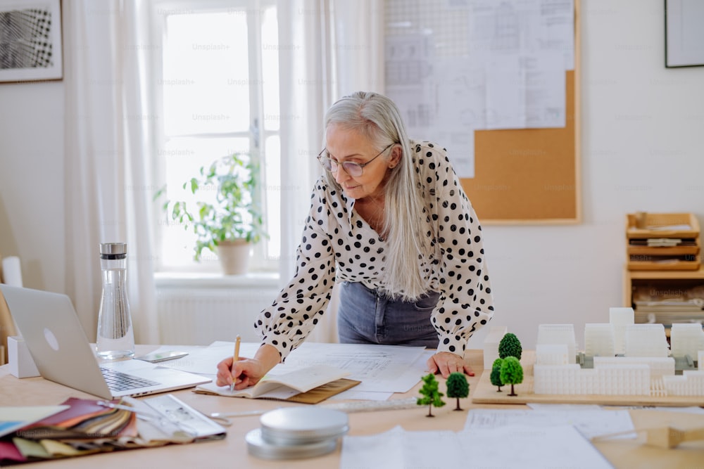 Un architetto donna anziana con un modello di case in piedi in ufficio e che scrive appunti.