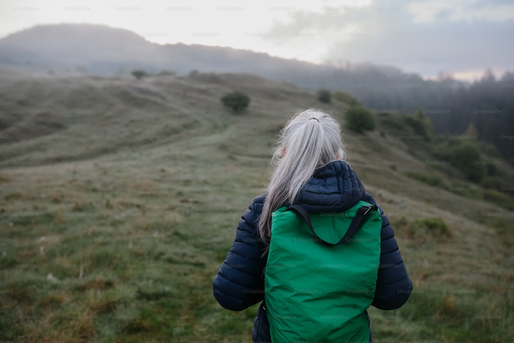 Uma vista traseira da mulher idosa caminhando na natureza no início da manhã com neblina e montanhas ao fundo.