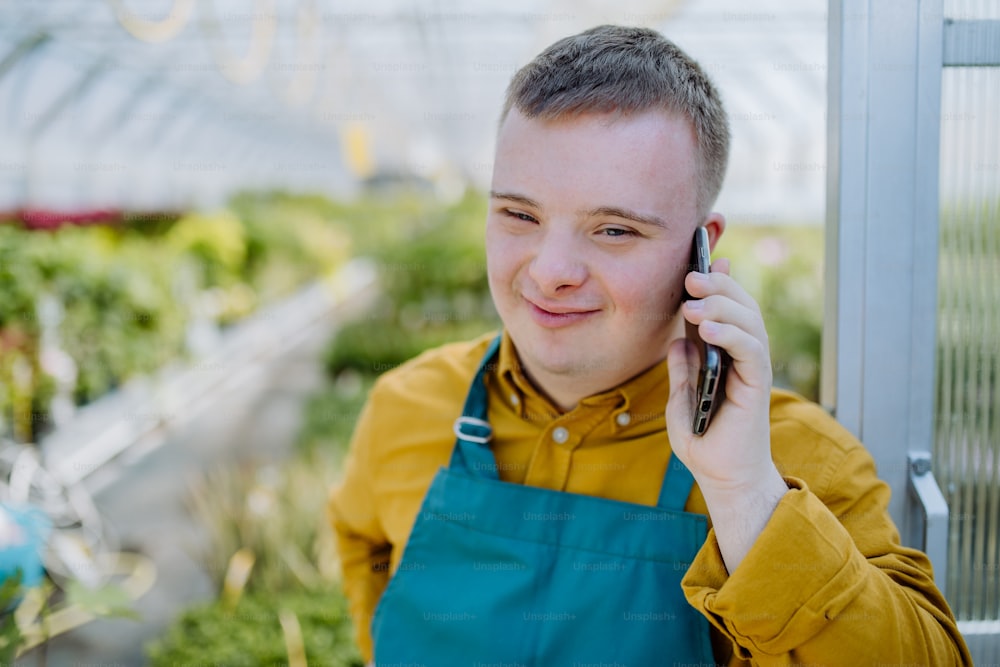 Un joven empleado con síndrome de Down que trabaja en un centro de jardinería, parado en la puerta del invernadero y llamando por teléfono celular.