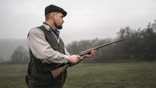 A hunter man in traditional shooting clothes on field aiming with shotgun.