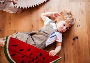 A top view of toddler boy playing with large toy fruit indoors at home, lying on the floor.