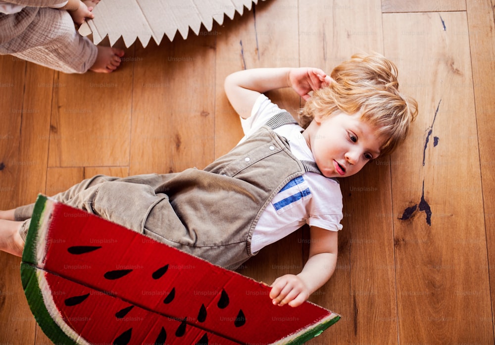A top view of toddler boy playing with large toy fruit indoors at home, lying on the floor.