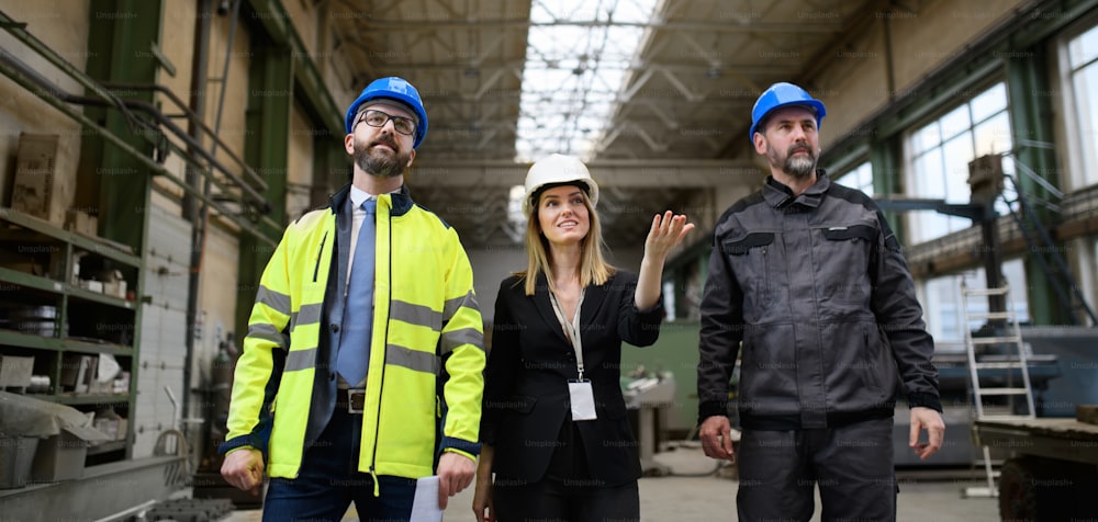 Manager supervisors and industrial worker in a uniform walking in large metal factory hall and talking. Low angle view.