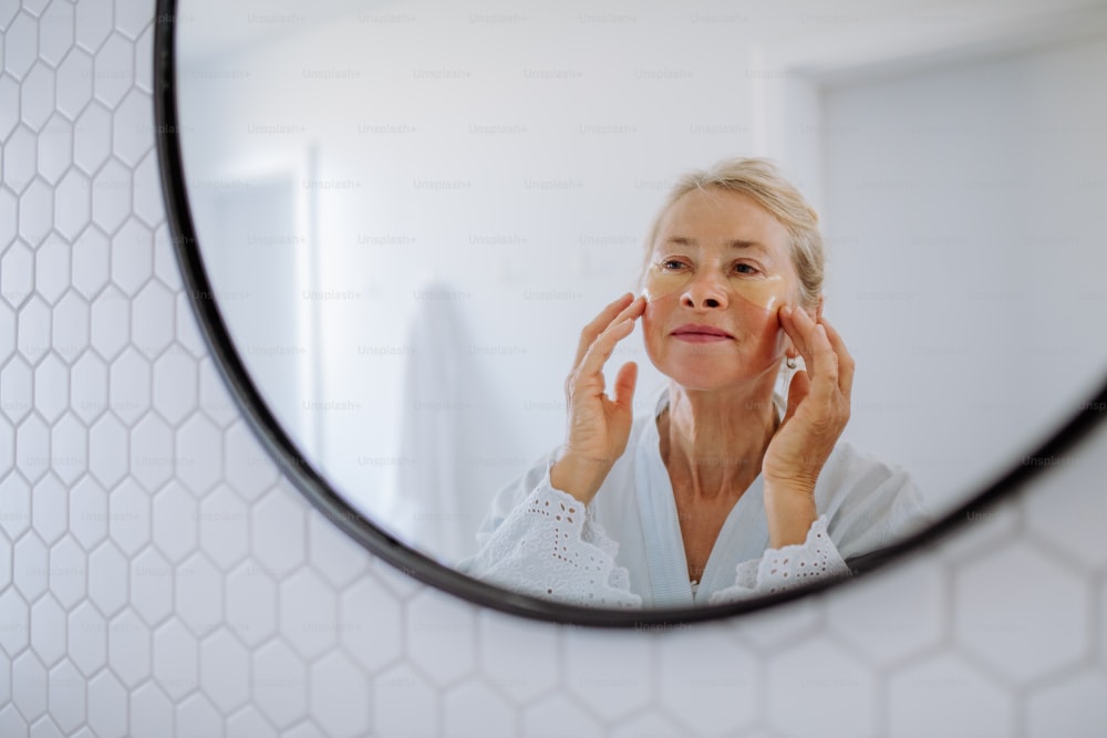 Una hermosa mujer mayor en bata de baño, aplicando parches en los ojos para la hinchazón mientras se mira en el espejo