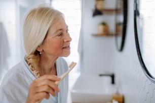 Une femme âgée en peignoir se brossant les dents avec une brosse à dents en bois écologique dans la salle de bain, un mode de vie durable.