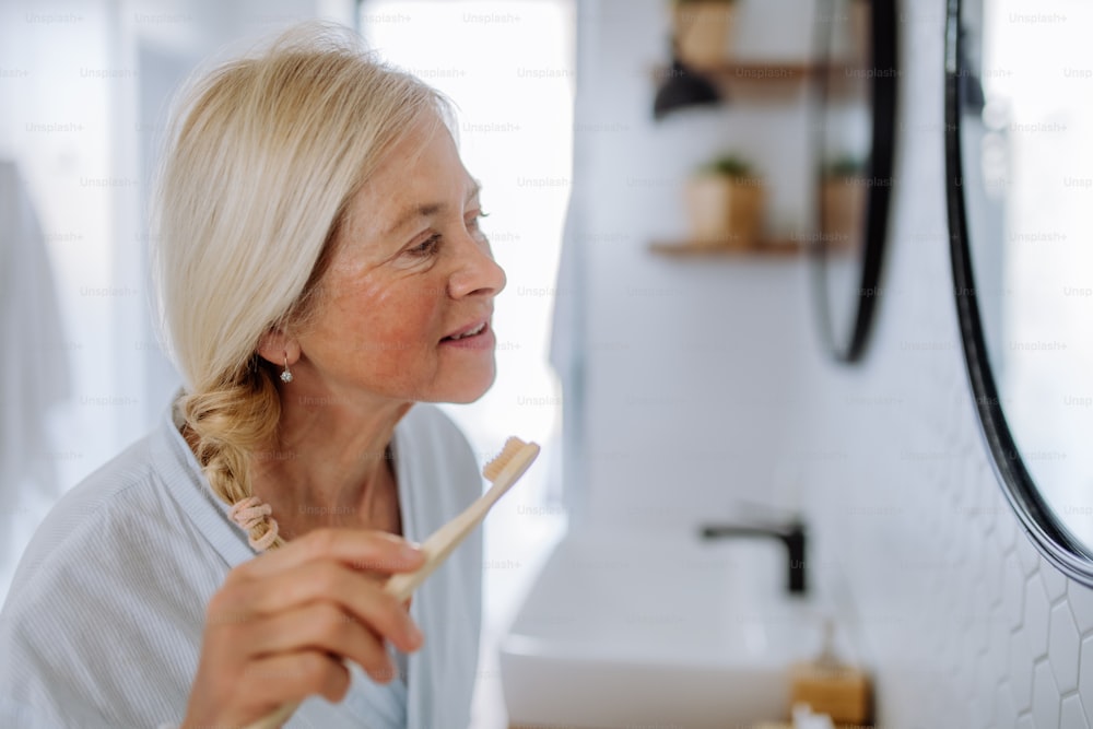 バスローブを着た年配の女性が、バスルームでエコな木製歯ブラシで歯を磨く、持続可能なライフスタイル。