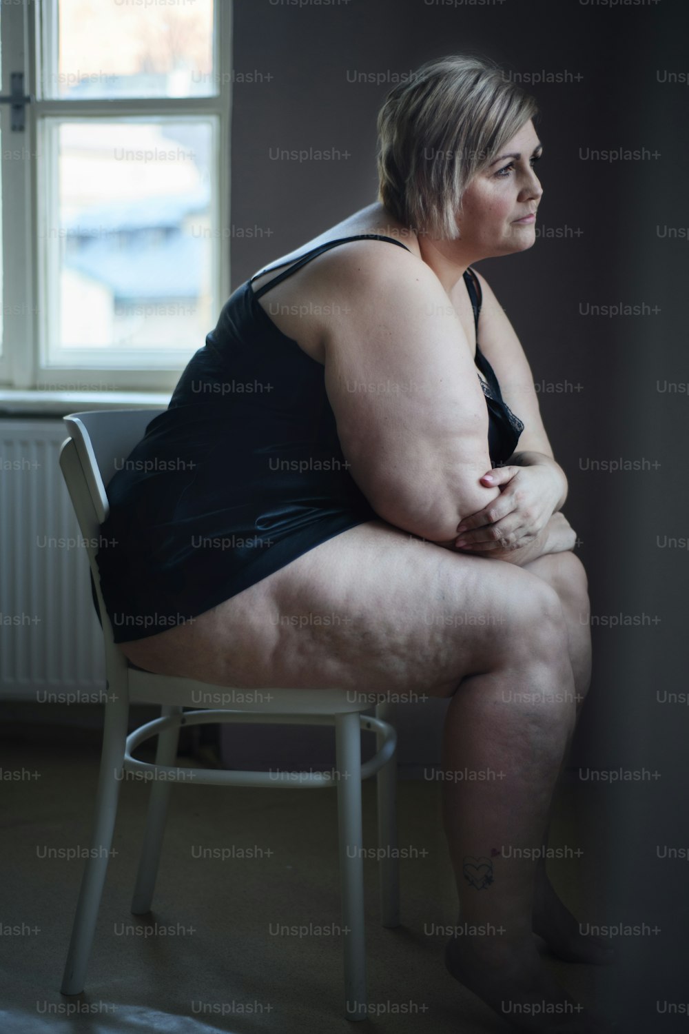 Depressed lonely fat woman sitting and looking through the window at home at dark.