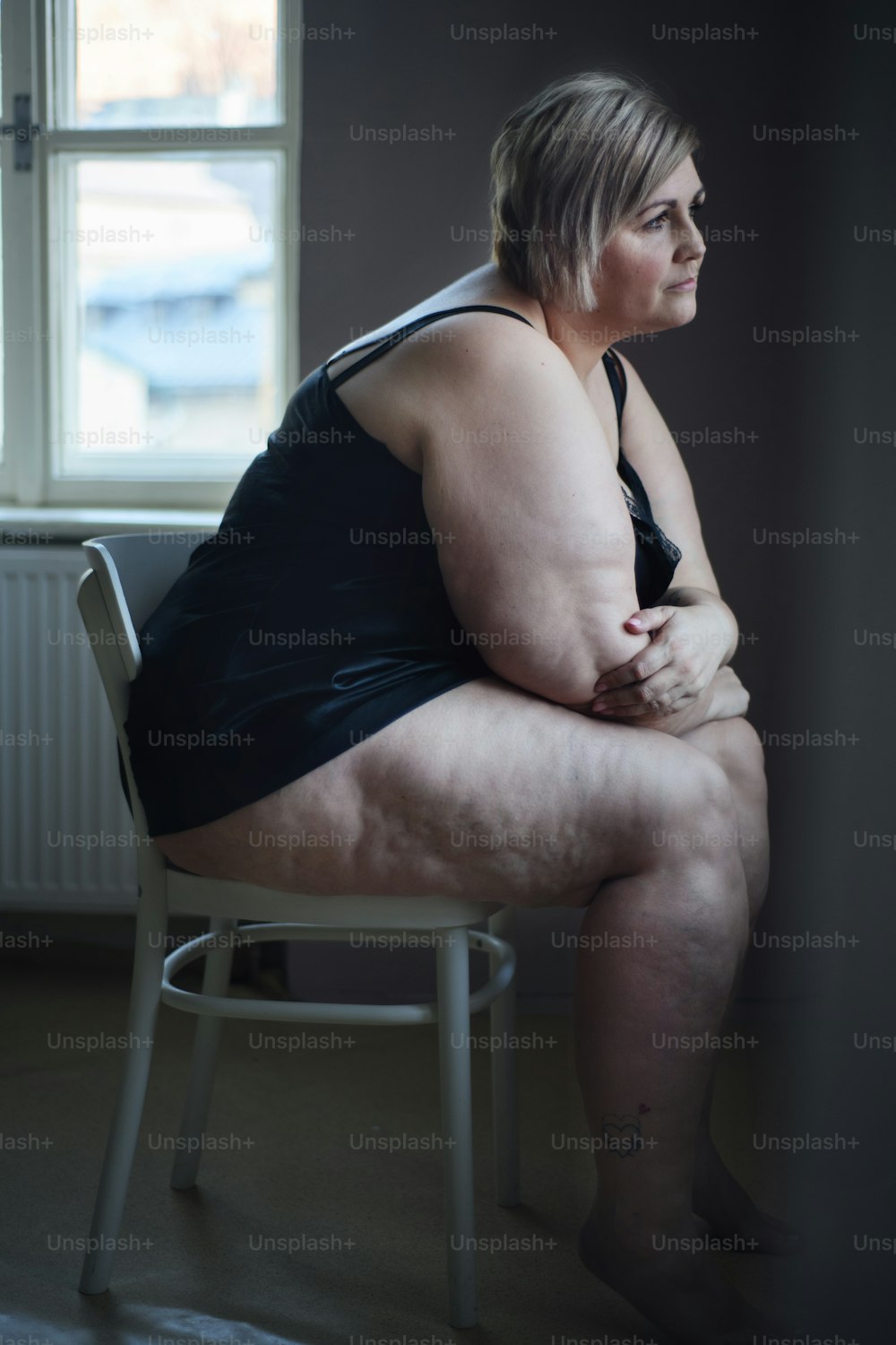 Deprimierte einsame dicke Frau, die im Einbruch der Dunkelheit zu Hause sitzt und durch das Fenster schaut.