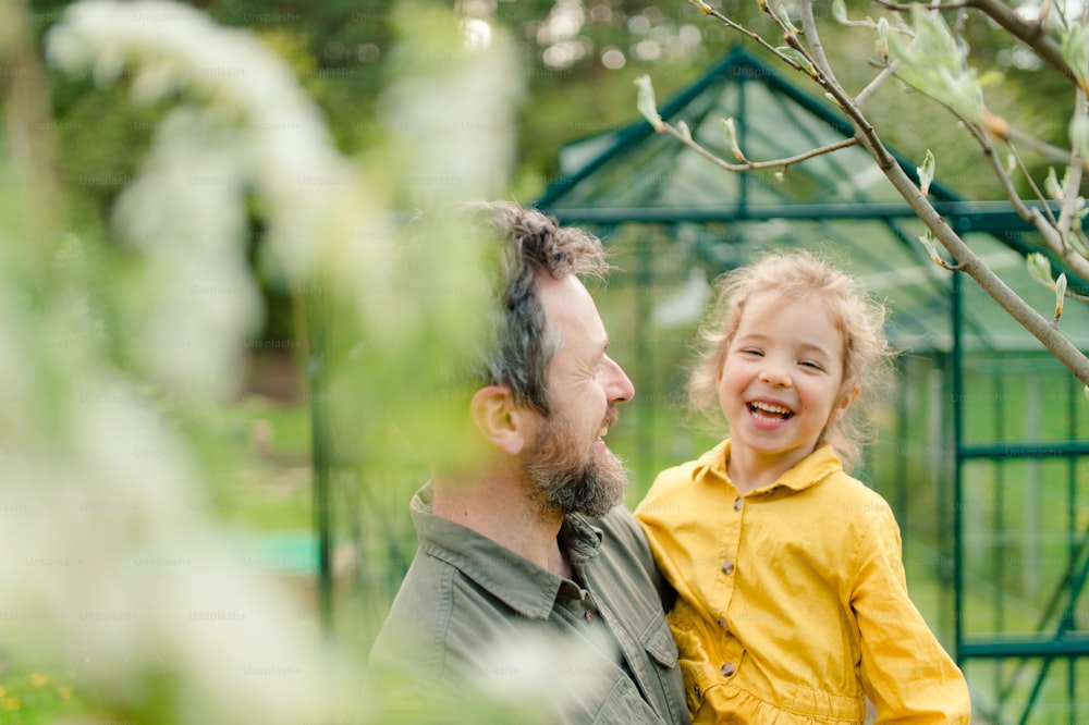 Un père avec sa petite fille riant devant une serre écologique, un mode de vie durable.