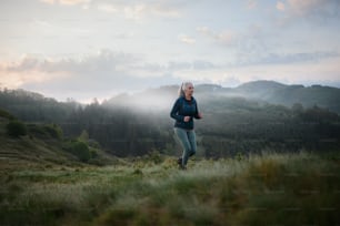 A senior woman jogging in nature on early morning with fog and mountains in background.