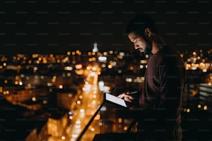 Un jeune homme afro-américain debout sur un balcon avec vue sur la ville et utilisant une tablette la nuit