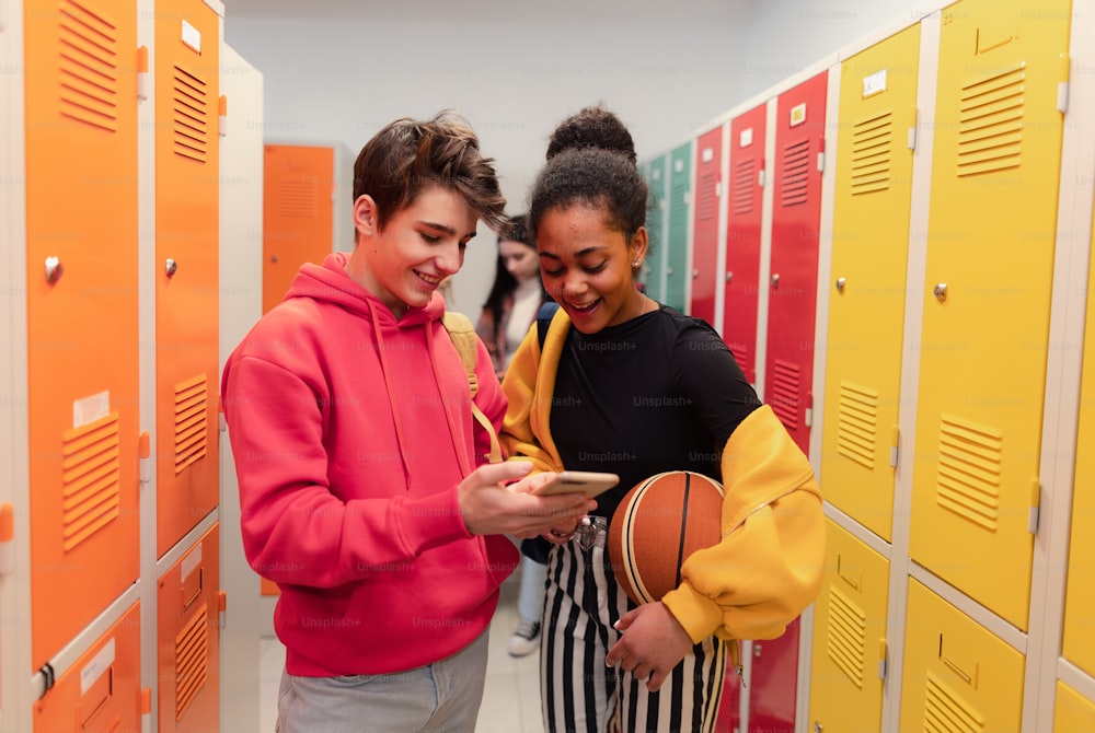 Young high school students standing near locker in campus hallway talking and using a smartphone.