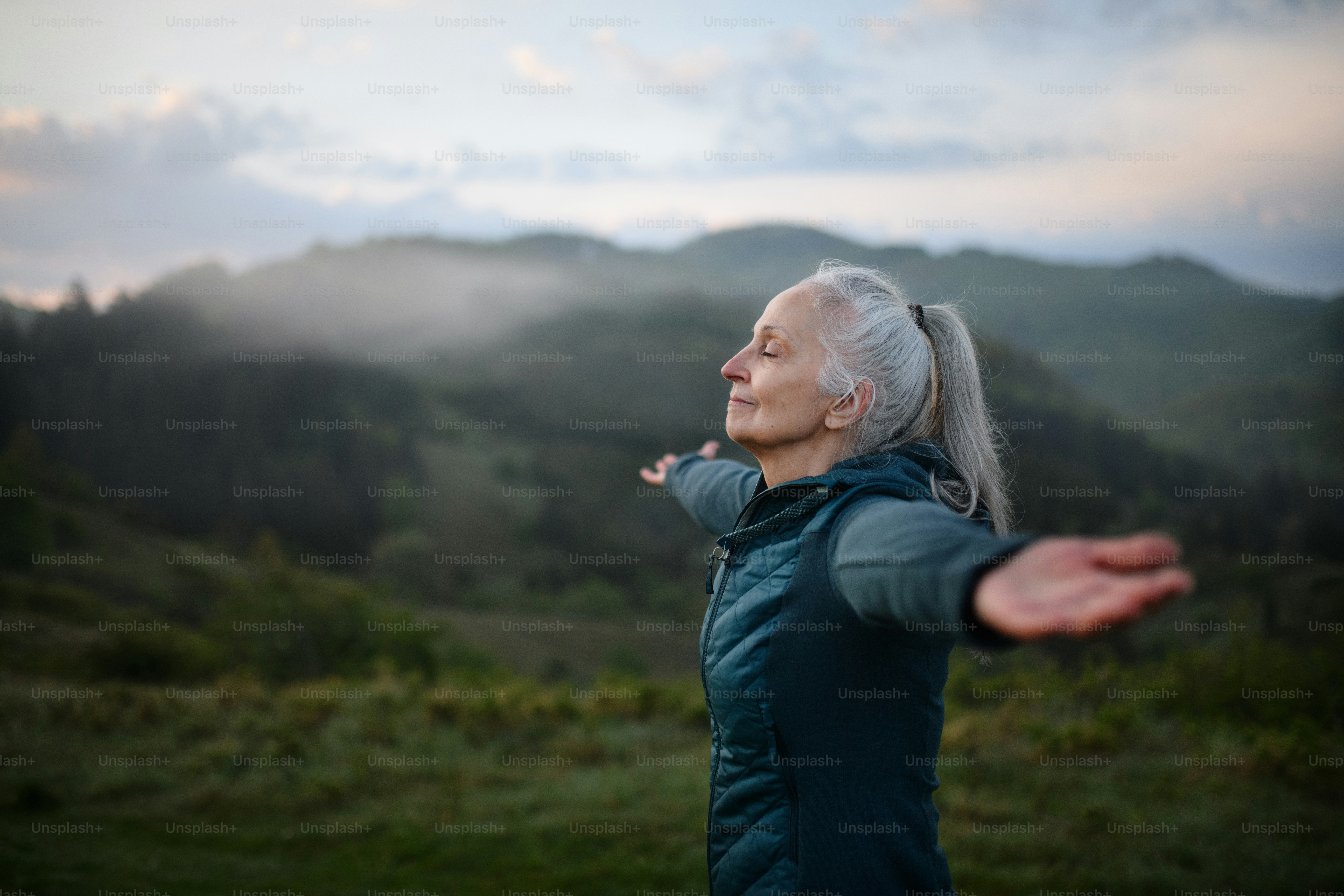 Une femme âgée faisant des exercices de respiration dans la nature tôt le matin avec du brouillard et des montagnes en arrière-plan.